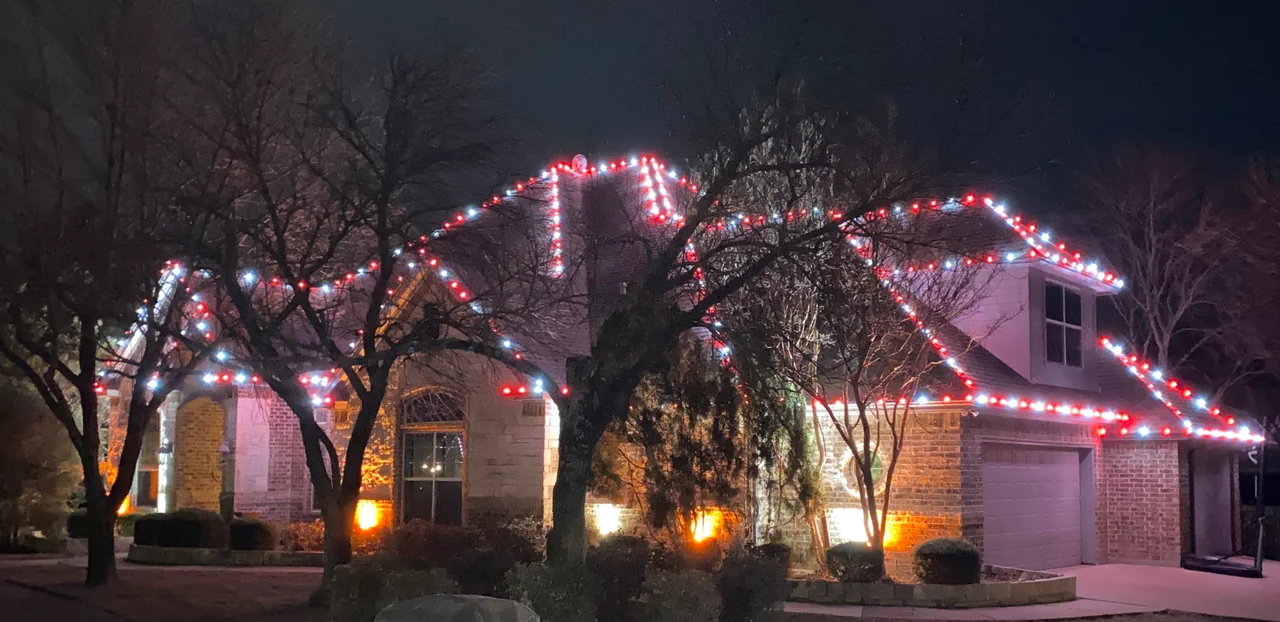 Red-and-white Christmas lights trace a two-story home behind mature trees.