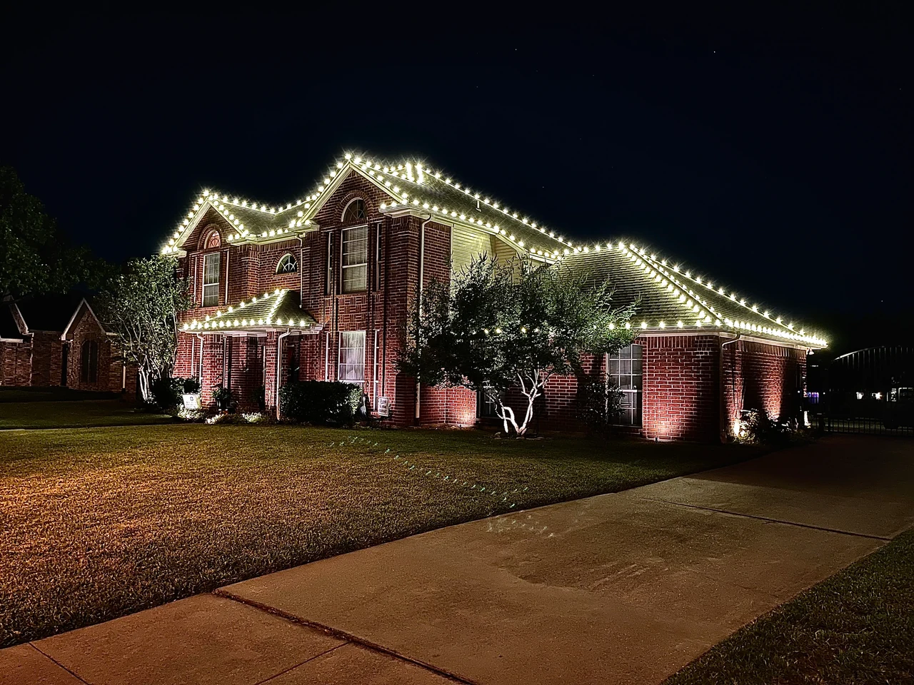 Warm-white Christmas lights outline the rooflines and peaks of a large two-story brick home.