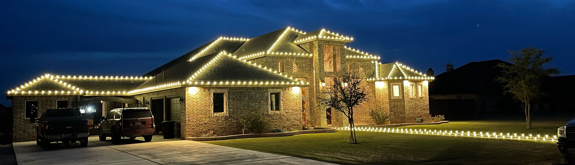 Warm-white Christmas lights trace the peaks and rooflines of a large brick home at blue hour.